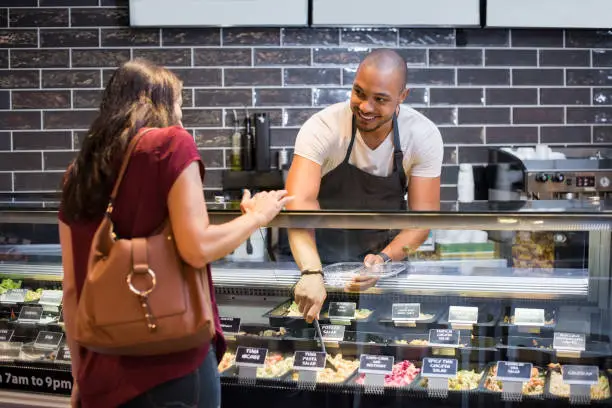 African waiter serving fresh food to young woman. Happy smiling guy preparing take away salad for woman customer. Black man with apron taking pasta salad in spoon and serving to customer.