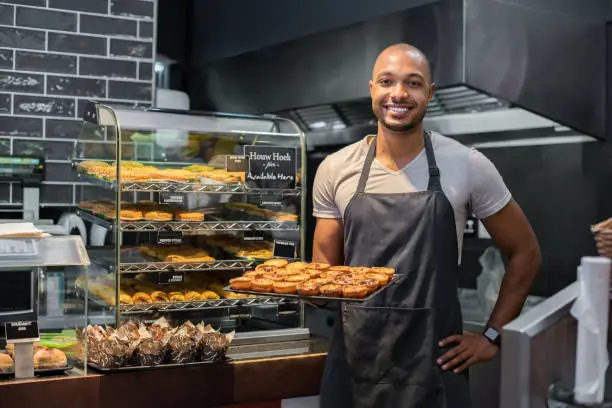 Smiling black baker with apron holding tray of small pastry and looking at camera. Young african chef holding sweet tray at cafeteria. Happy black man smiling at bakery.
