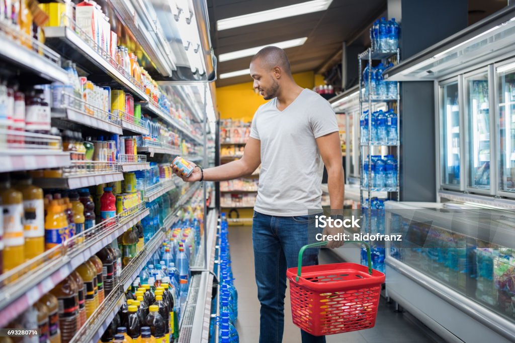 Man at supermarket African man shopping in beverage section at supermarket. Black man doing shopping at market while buying cold drink. Handsome guy holding shopping basket reading nutritional values of product. Supermarket Stock Photo