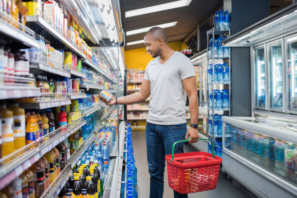 Man at supermarket African man shopping in beverage section at supermarket. Black man doing shopping at market while buying cold drink. Handsome guy holding shopping basket reading nutritional values of product. grocery shelf cans stock pictures, royalty-free photos & images