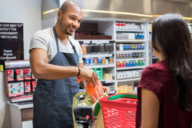 Cashier working at supermarket Smiling salesman putting vegetables in bag for customer after billing. Cashier black man at grocery store helping customer pack purchased products. Happy young man working in grocery shop. cashier stock pictures, royalty-free photos & images