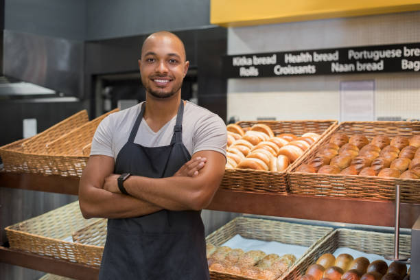 Happy african baker man Smiling baker man standing with fresh bread at bakery. Happy african man standing with crossed arms at counter in bake shop and looking at camera. Satisfied baker with breads in background. baker occupation stock pictures, royalty-free photos & images