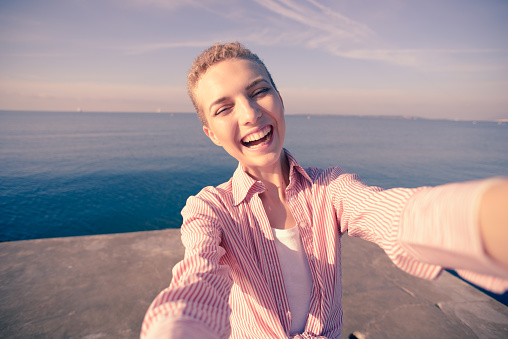 Camera point of view - happy laughing young woman uses a smart phone to take a selfie, film a video, or make a video call; standing on a jetty in the sea at dawn in the sunshine, Antibes Juan-les-Pins, Cote d'Azur, France.