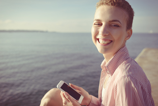 Happy laughing young woman uses a smart phone app or makes a video call; sitting on a jetty in the sea at dawn in the sunshine, Antibes Juan-les-Pins, Cote d'Azur, France.