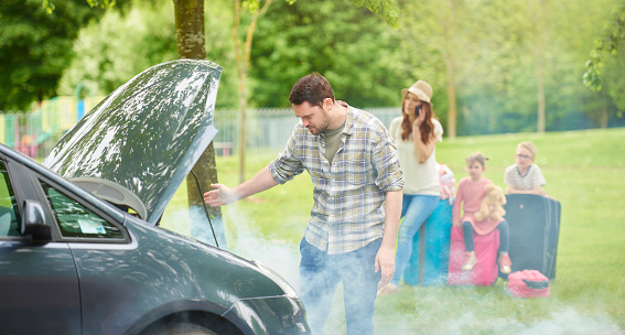 a father assesses the problem with his broken down car as his wife sits roadside with the luggage calling the breakdown truck.takes the children off to school in the morning