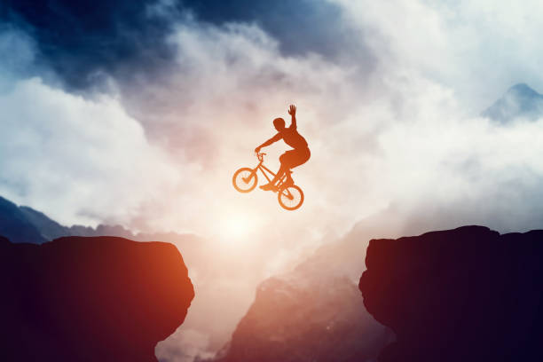 Man jumping on bmx bike over precipice in mountains at sunset. Man jumping on bmx bike over precipice in mountains at sunset. Raising hand showing hello gesture. Extreme sport, risk, cycling. bicycle stunts stock pictures, royalty-free photos & images