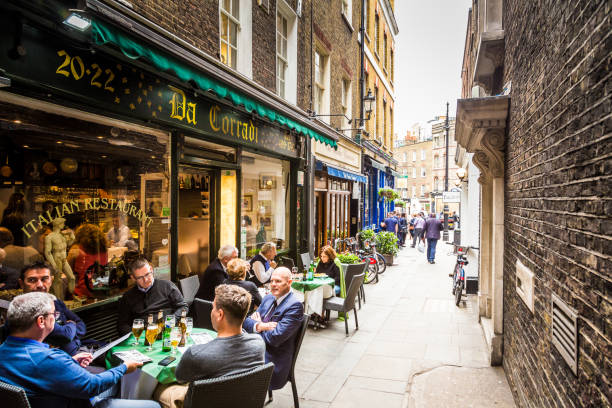 Business people eating lunch outdoors in Mayfair, central London, UK London, UK - May 18, 2017: businessmen enjoy an al fresco lunch at an Italian restaurant in Mayfair, an exclusive, wealthy area in central London, UK. The men are drinking beers and looking at the menu. The street is narrow and quaint, emphasising the diminishing perspective of the image. Horizontal colour image with copy space. Mayfair stock pictures, royalty-free photos & images