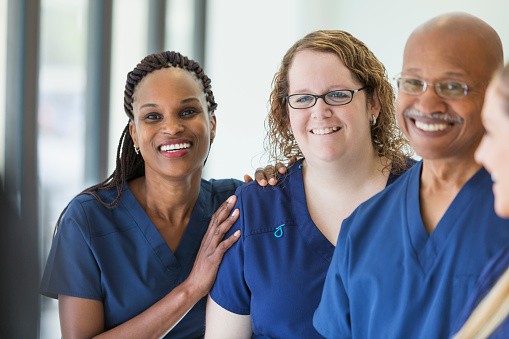 A team of multi-ethnic medical professionals standing in a corridor, wearing blue scrubs, smiling. The focus is on the two women. The African-American woman is in her 40s and the one wearing eyeglasses is in her 30s.