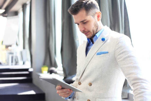 istock Working on new tablet. Low angle view of confident young man working on digital tablet while standing in front of the big window in office or cafe 693439432