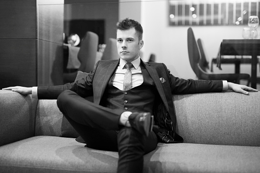 Black and white fashion portrait of a handsome young man sitting on the sofa at the hotel lobby.