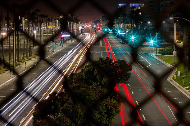 a vivid night shot of  the Pacific Coast Highway (PCH) in Huntington Beach, California.  Creatively Shot through the Chain Link fence of a pedestrian bridge over PCH  This features a long exposure of taillights and headlights, on a vivid background of Downtown Huntington Beach.