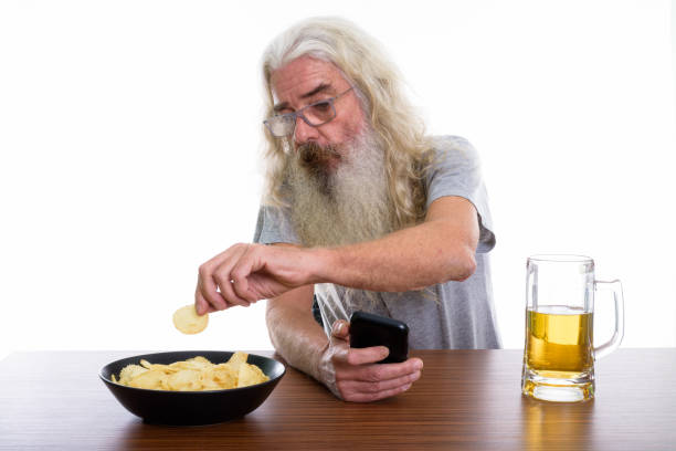 木製のテーブルにビールとポテトチップスのボウルを食べながら携帯電話を保持しているシニアのひげを生やした男のスタジオ撮影 - beer glass mustache beer color image ストックフォトと画像