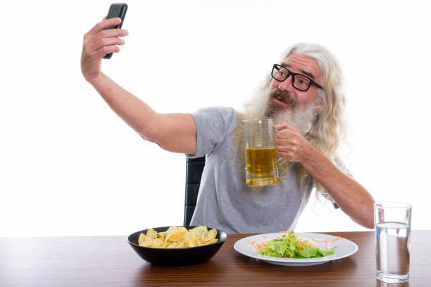 homem barbudo sênior feliz sorrindo enquanto estiver a tomar selfie foto com o telemóvel com o copo de cerveja com alimentos e não-íntegros na mesa de madeira - beer glass mustache beer color image - fotografias e filmes do acervo