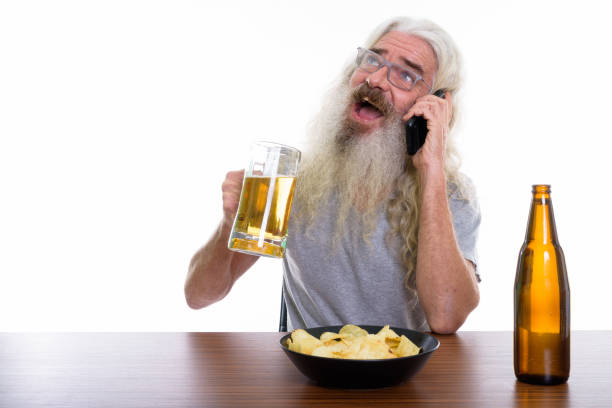 pensativo sênior barbudo homem feliz sorrindo enquanto falando no telefone móvel e segurando o copo de cerveja com o prato de batatas fritas na mesa de madeira - beer glass mustache beer color image - fotografias e filmes do acervo