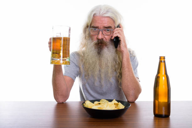 foto de estúdio de sênior homem barbudo falando no celular com copo de cerveja com o prato de batatas fritas na mesa de madeira - beer glass mustache beer color image - fotografias e filmes do acervo