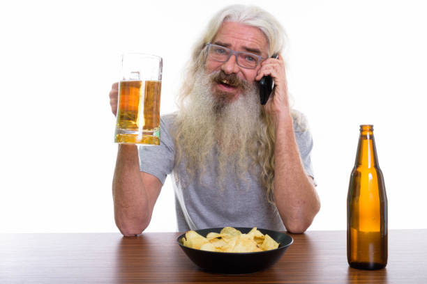 foto de estúdio do homem barbudo sênior feliz sorrindo e falando no celular com copo de cerveja com o prato de batatas fritas na mesa de madeira - beer glass mustache beer color image - fotografias e filmes do acervo