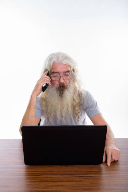 istock Studio shot of senior bearded man wearing eyeglasses and talking on mobile phone while using laptop on wooden table 693579902