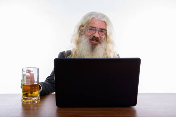studio um shot de feliz empresário barbudo sênior, sorrindo enquanto usando laptop com copo de cerveja na mesa de madeira - beer glass mustache beer color image - fotografias e filmes do acervo