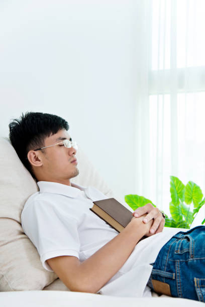 istock Young asian man with book asleep on sofa 693610912