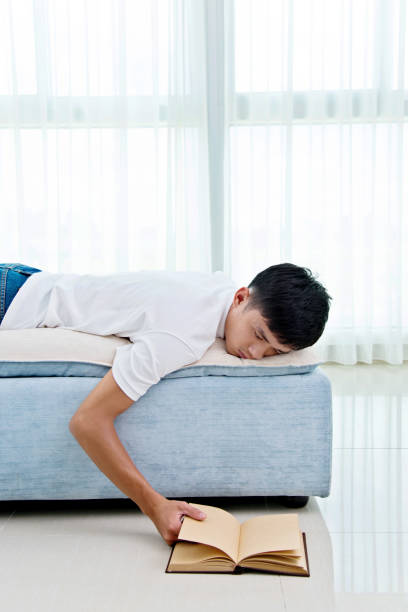 istock Young asian man with book asleep on sofa 693611086