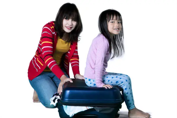 Portrait of Asian mother and her daughter ready to vacation while trying to close their suitcase, isolated on white background