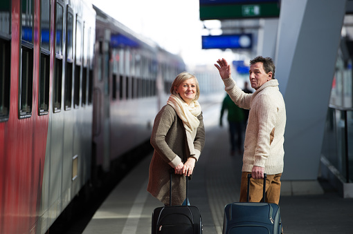 Beautiful senior couple on trainstation pulling a trolley luggage, waving.
