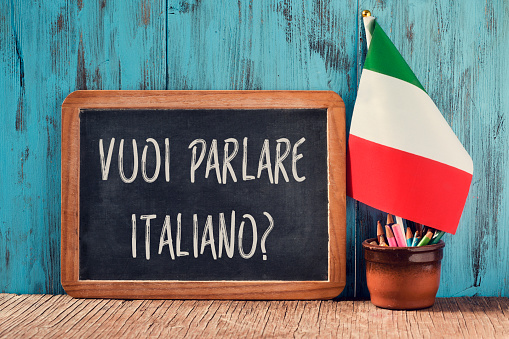 a chalkboard with the question vuoi parlare italiano?, do you want to speak Italian? written in Italian, a pot with pencils, some books and the flag of Italy on a wooden desk