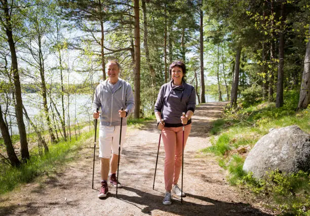 Summer sport in Finland - nordic walking. Man and mature woman hiking in green sunny forest. Active people outdoors. Scenic peaceful Finnish summer landscape.