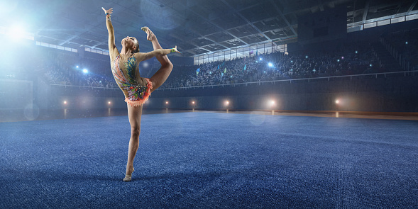 A gymnast girl makes performance on a large professional stage. She is wearing a professional swimsuit for gymnastics. Behind her is a stadium with spectators.