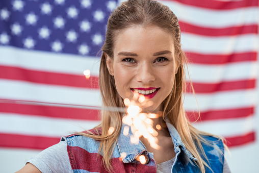 portrait of happy woman holding sparkler with American flag behind, Independence Day Celebration