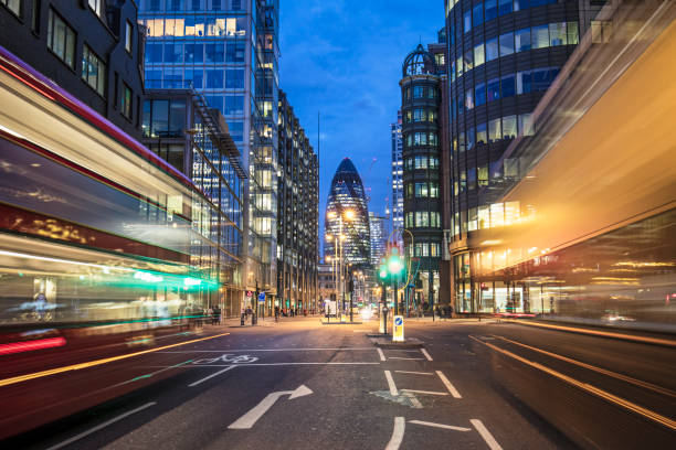 Vehicles moving on street against 30 St Mary Axe Blurred motion of vehicles moving on street with 30 St Mary Axe in background. Illuminated towers are in city. View of downtown district at night. london gherkin at night stock pictures, royalty-free photos & images