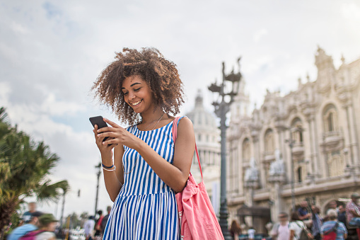 Young woman texting on smart phone against National Theater of Cuba. Beautiful female is smiling while traveling in city. She is in striped blue dress.