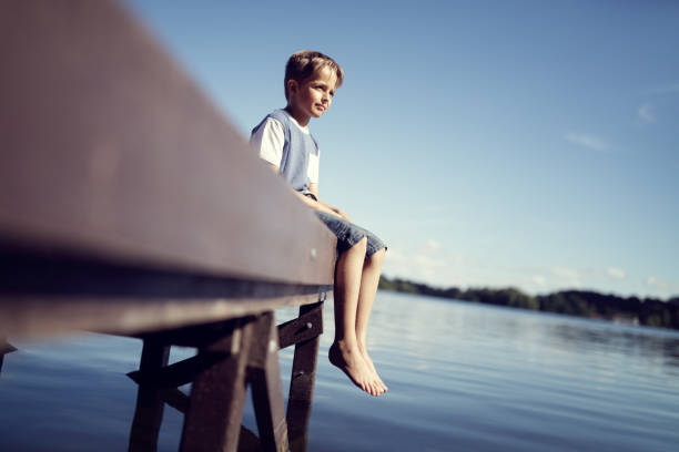 boy with legs dangling from pier - beira dágua imagens e fotografias de stock