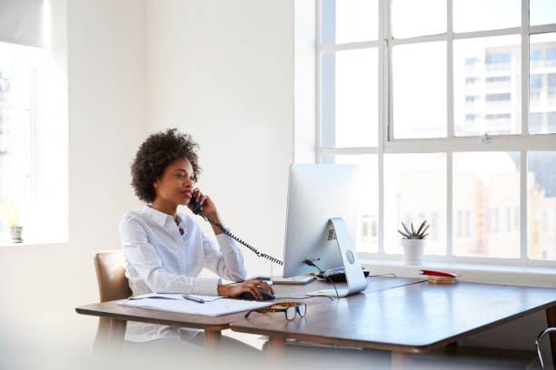 Young black woman talking on phone at her desk in an office Young black woman talking on phone at her desk in an office landline phone stock pictures, royalty-free photos & images