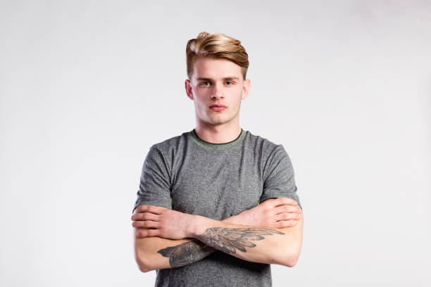 Handsome man in gray t-shirt, tattoo on forearms, studio shot. Young handsome hipster man in gray t-shirt, tattoo on his forearms, arms crossed. Studio shot on gray background. arm tattoo ideas for men pictures stock pictures, royalty-free photos & images