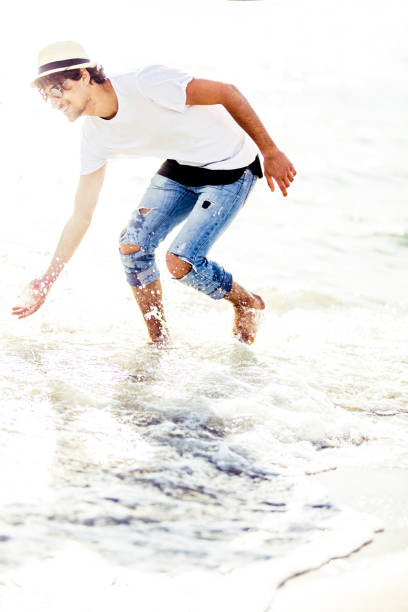 Young beautiful and fashionable hipster man walking on the beach near the water and enjoying the summer day