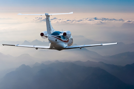 Private jet, passenger wide-body plane or aircraft is flying in the blue sky over the the clouds and mountains. Summer vacation concept.