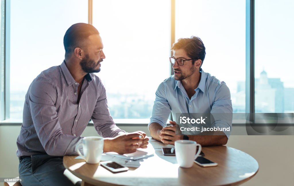 Business partners discussing business plans sitting at table in office. Business executives discussing work in office. Two businessmen sitting on a round table and discussing business matters. Discussion Stock Photo