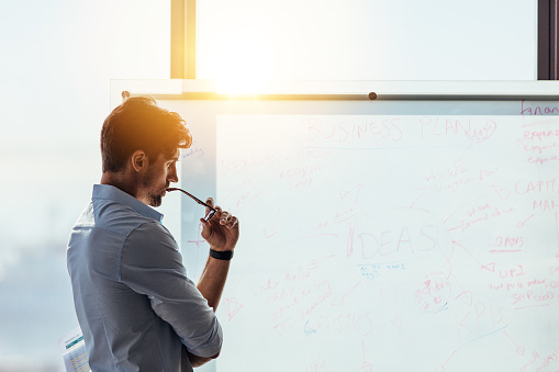 Business investor in deep thought looking at the business ideas written on the whiteboard. Businessman thinking while holding his spectacles to mouth