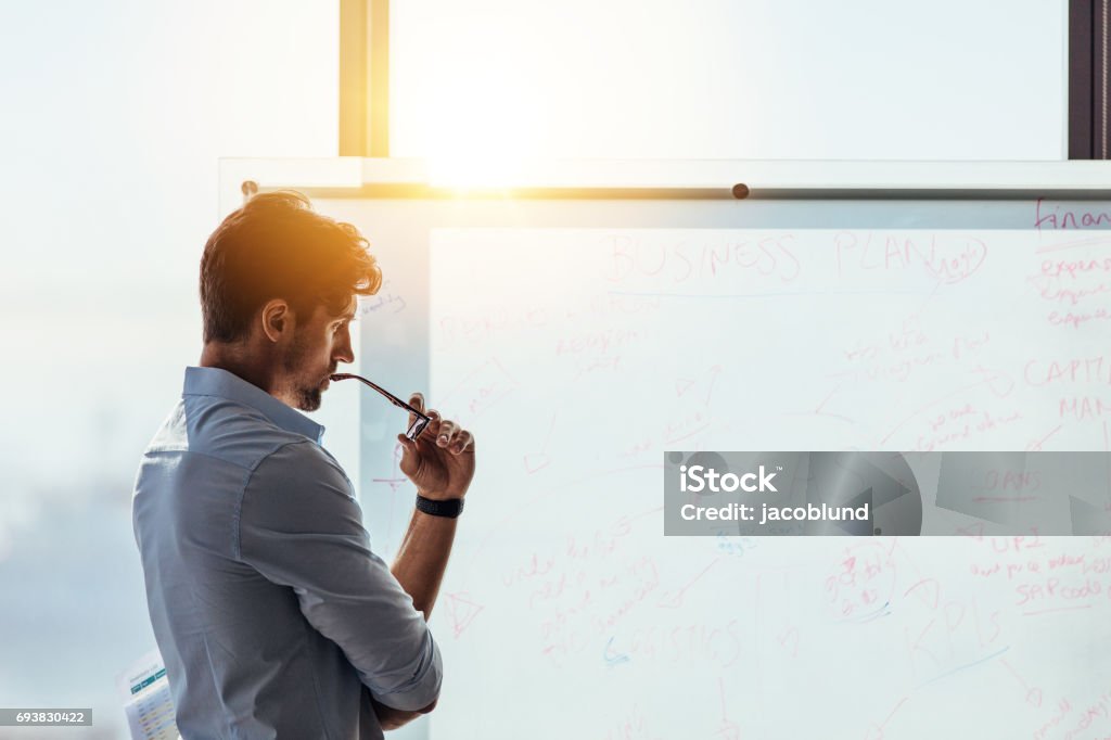 Entrepreneur putting his business ideas on whiteboard in boardroom. Business investor in deep thought looking at the business ideas written on the whiteboard. Businessman thinking while holding his spectacles to mouth Contemplation Stock Photo