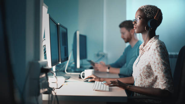 Graphic designers at work. Closeup side view of mid 20's blond black woman doing her design project on a computer. She might be a software developer as well.  She's sitting in front of a desktop computer and sipping a coffee while working on a computer. outsourcing stock pictures, royalty-free photos & images