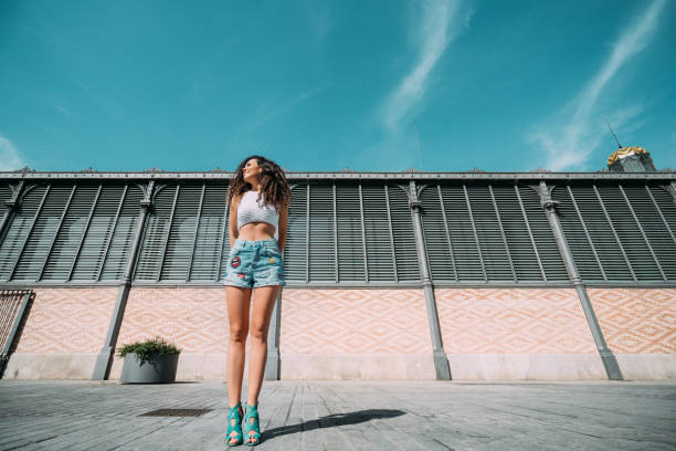 Smiling hipster girl standing on street with long building in background with copy space for young advertising text message, curly happy slim brunette woman looking outside on sunny day with teal sky