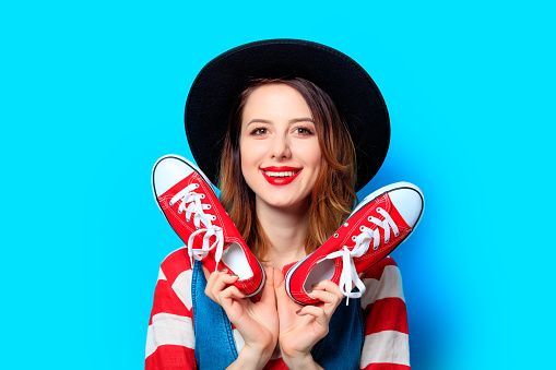 Portrait of young smiling red-haired white european woman in hat and red striped shirt with gumshoes ready for shopping on blue background