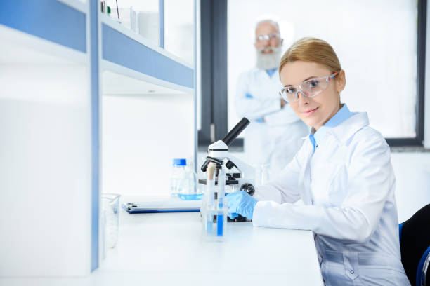 istock Smiling middle aged scientist in protective eyewear working with microscope and colleague standing behind 693992446