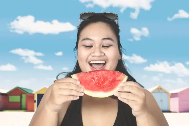 Fat woman wearing swimsuit, eating a slice of watermelon near the cottage