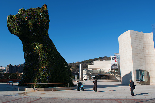 Bilbao, Spain - January, 26, 2017: people at the Guggenheim Museum Bilbao, the museum of modern and contemporary art designed by architect Frank Gehry, opened in 1997, among the most admired works of contemporary architecture, and view of the flower sculpture Puppy, created in 1992 by the artist Jeff Koons