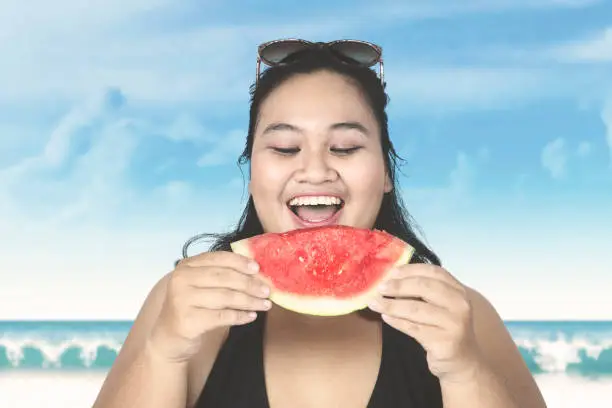 Obese woman wearing swimsuit, eating fresh watermelon on the beach