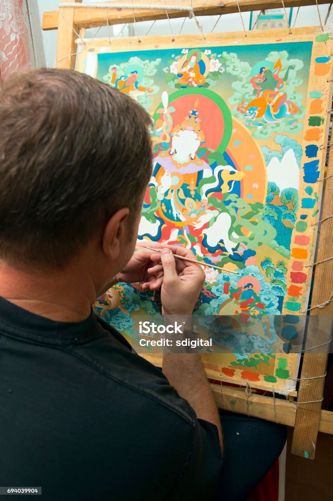 The artist paints a Buddhist icon The artist sitting cross-legged in the lotus pose draws a Buddhist icon of the tank with a thin column brush in the art workshop. Close-up of the hand of the draftsman with a brush and a palette Paint Stock Photo