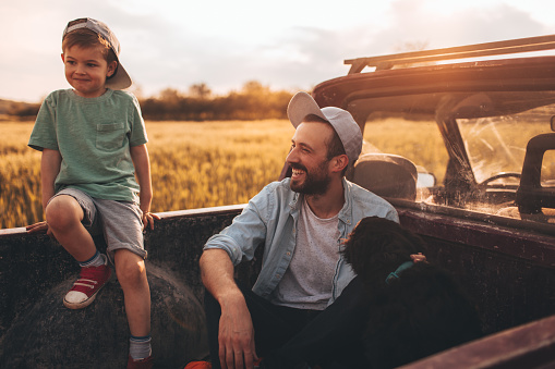 Photo of young father bonding with his boy during the ride in a family pick-up truck, outdoors in the nature. They are in a company of a family dog.