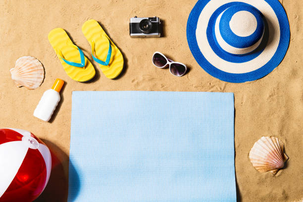 Summer vacation composition. Sandals, hat and beach ball. Summer vacation composition with pair of yellow flip flop sandals, hat, sunglasses, sun screen, beach ball and blue mat on a beach. Sand background, studio shot, flat lay, copy space. beach mat stock pictures, royalty-free photos & images